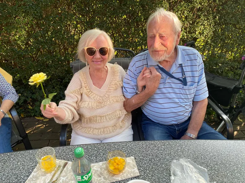 Eine Frau und ein Mann sitzen händchenhaltend an einem Tisch. Die Frau hält eine Blume in ihrer anderen Hand