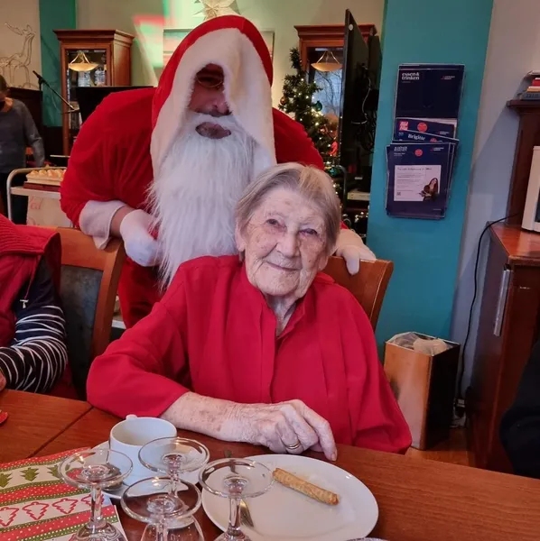 eine alte Frau sitzt an einem Tisch und lächelt in die Kamera. Hinter ihr steht ein verkleideter Weihnachtsmann