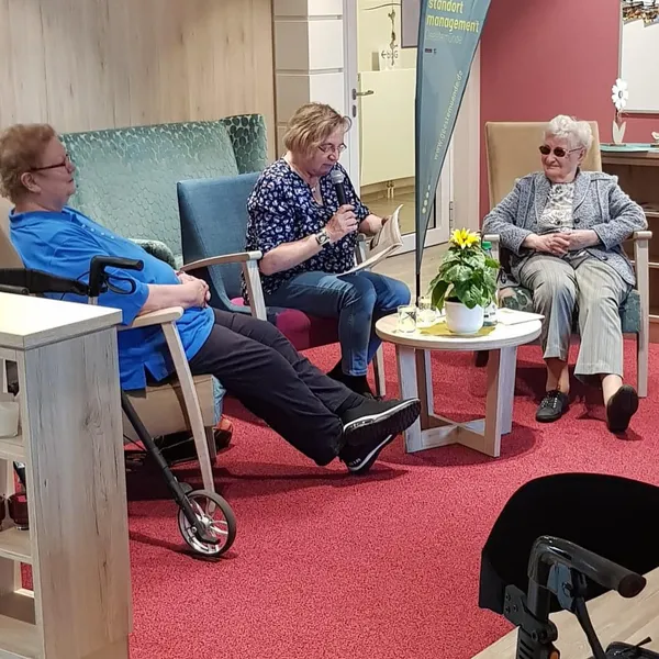 Drei Frauen sitzen um einen kleinen Beistellstisch. Die Frau in der Mitte hat ein Mikrofon in der Hand und liest etwas vor