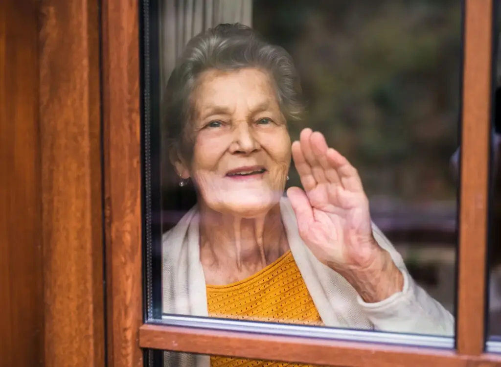 Eine Frau winkt durch ein Fenster der Kamera zu und lächelt freundlich