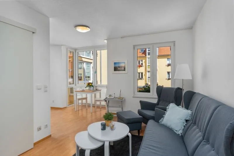 rechts ein dunkelgraues Sofa und Sessel mit Ottomanen, im Hintergrund ein kleiner Essbereich