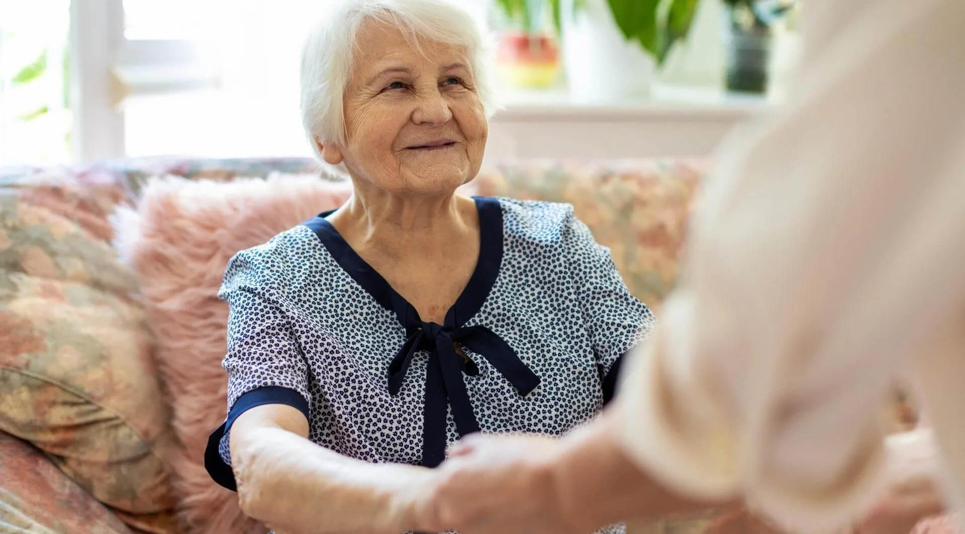 Alte Frau sitzt auf einem Sofa. Sie hält die Hände einer anderen Person und schaut diese lächelnd an