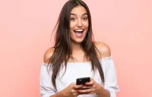 junge Frau schaut freudig in die Kamera mit weit aufgerissenen Mund und hat ein Handy in der Hand