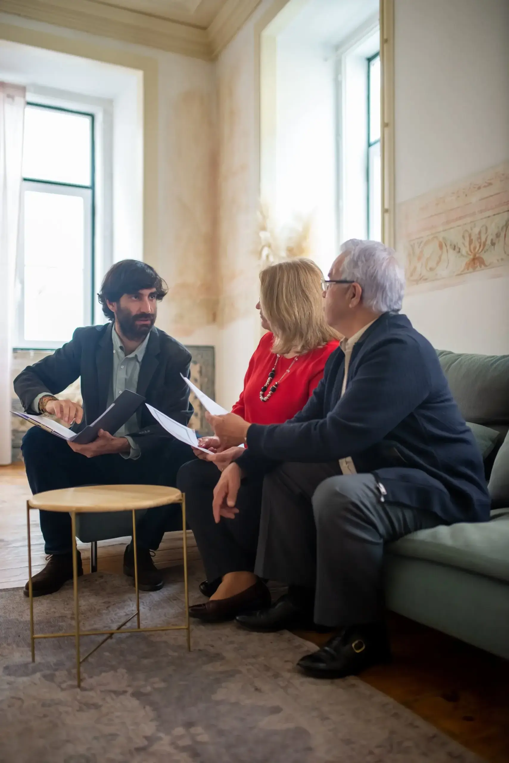 Rechts sitzen eine Frau und ein Mann auf einem Sofa. Daneben sitzt auf einem Hocker ein Mann mit einer Mappe in der Hand und erklärt etwas