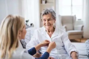 alte Frau sitzt auf einem Bett. eine zweite Frau mit dem rücken zur Kamera knöpft ihr das Hemd zu