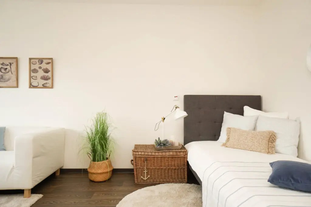 Blick auf ein Bett mit vielen Dekokissen und einer Ratterntruhe als Nachtschränkchen